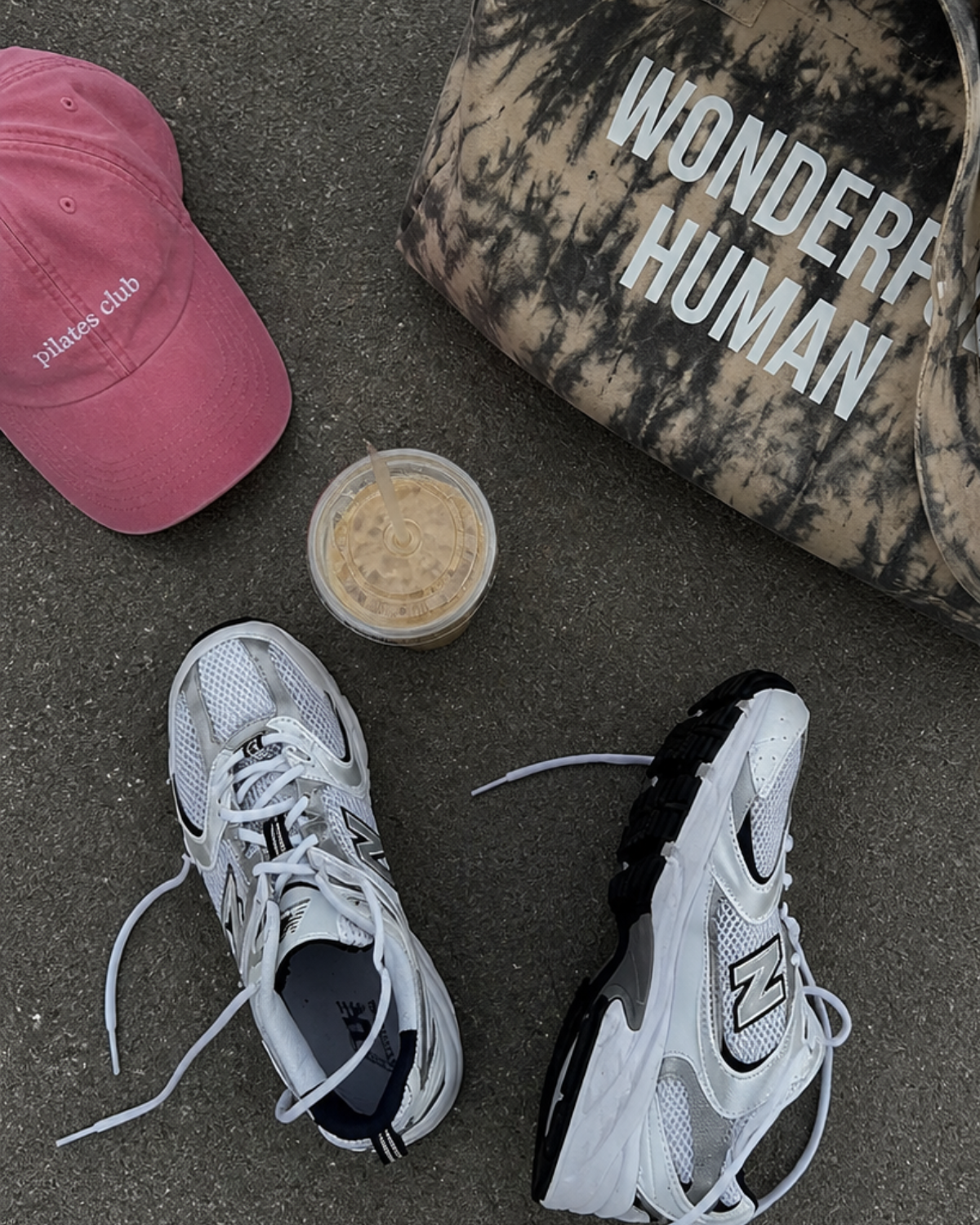 Two sneakers, a pink cap, a black and white cap with 'Wonder Human' text, and a small container on a concrete surface.