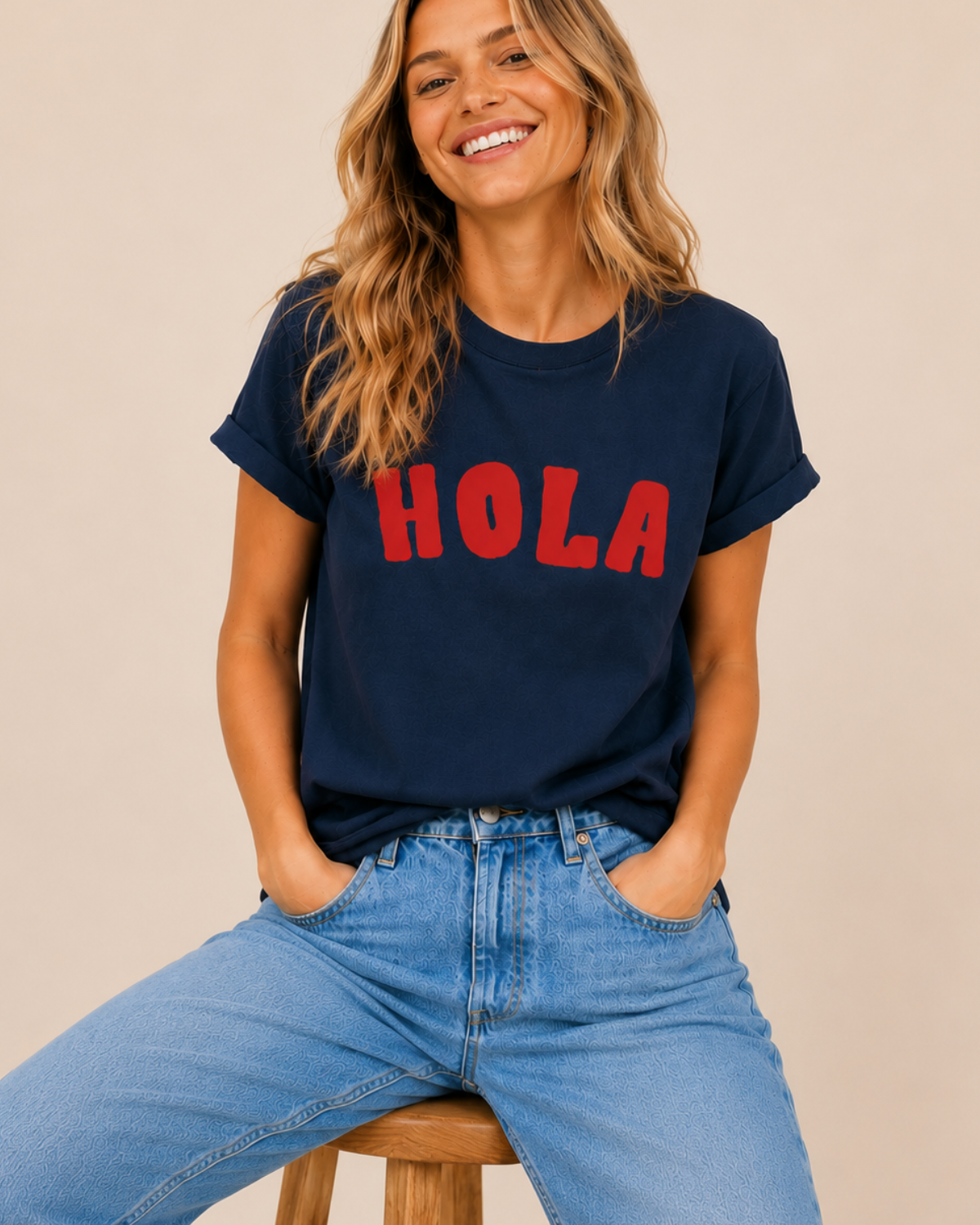 Woman wearing a navy t-shirt with 'HOLA' in red text and blue jeans, sitting on a wooden stool.