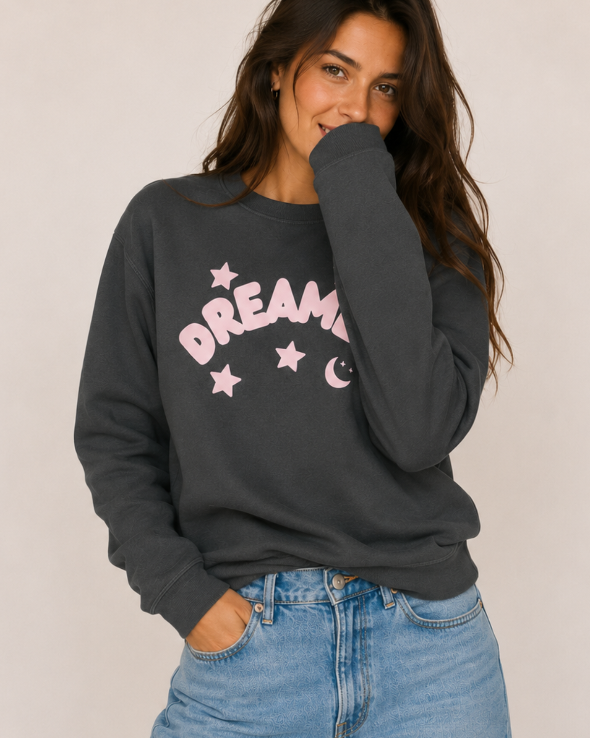 Woman wearing a dark gray sweatshirt with 'Dream' printed in pink and stars, paired with blue jeans on a plain background.