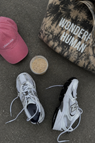 Two sneakers, a pink cap, a black and white cap with 'Wonder Human' text, and a small container on a concrete surface.