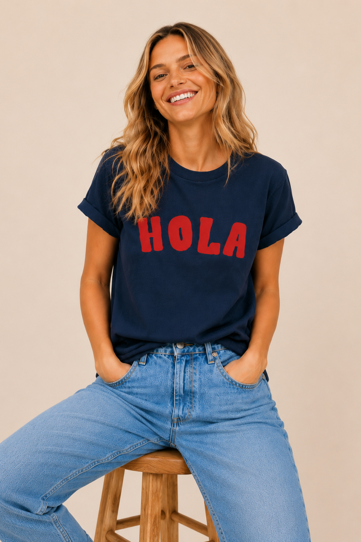 Woman wearing a navy t-shirt with 'HOLA' in red text and blue jeans, sitting on a wooden stool.