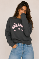 Woman wearing a dark gray sweatshirt with 'Dream' printed in pink and stars, paired with blue jeans on a plain background.